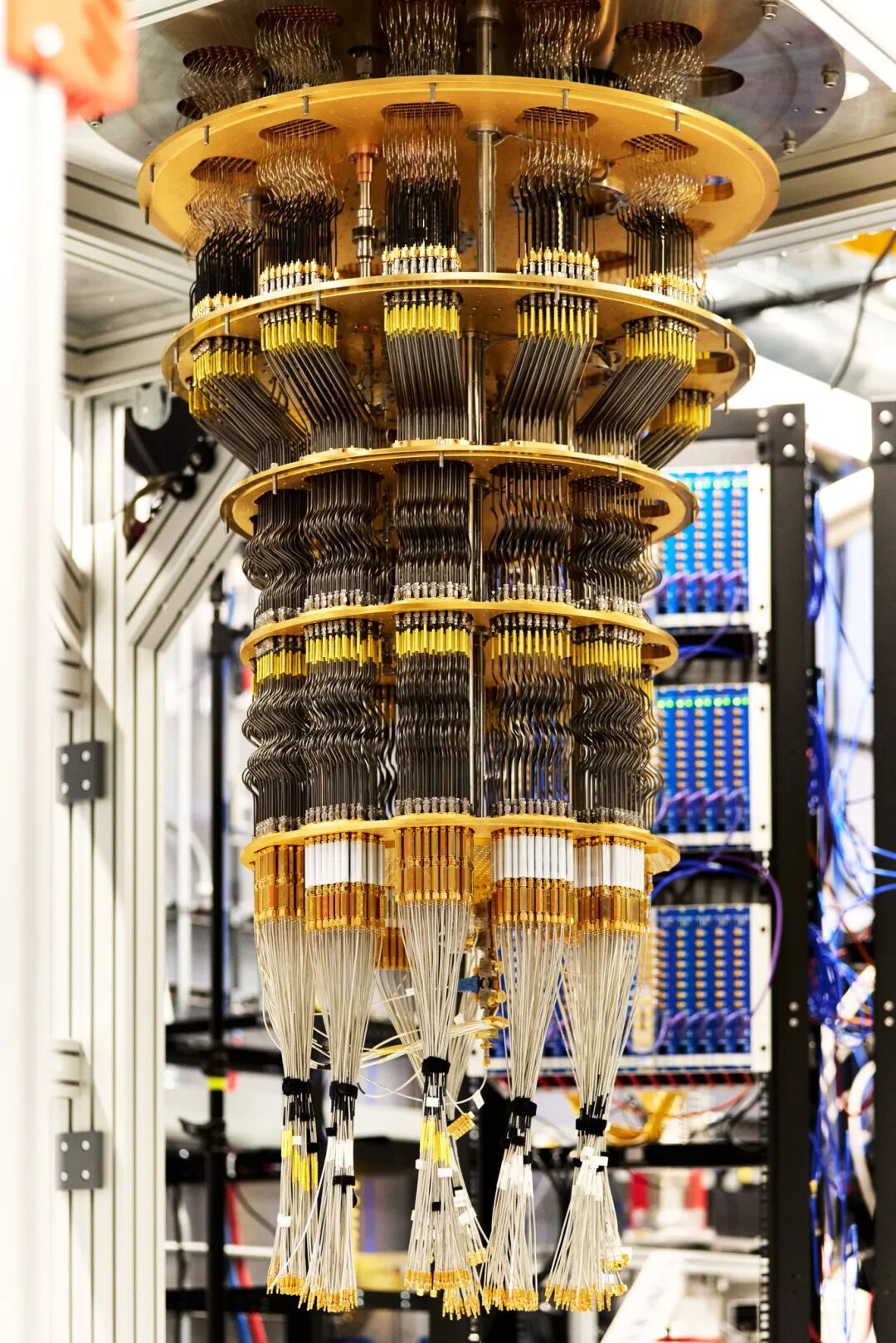 A complex cylindrical quantum chip assembly with multiple stacked layers of black and metallic components connected by numerous thin yellow and black cables extending downward surrounded by server racks and metallic framework in a laboratory setting